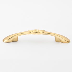 May include: A gold-toned cabinet pull with a decorative floral design. The pull is curved and has a smooth, polished finish.