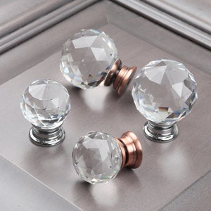 May include: Four clear crystal cabinet knobs with a faceted design. Each knob has a silver base and a copper colored ring around the base.
