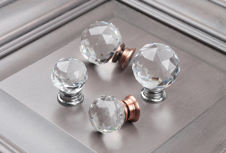 May include: Four clear crystal cabinet knobs with a faceted design. Each knob has a silver base and a copper colored ring around the base.