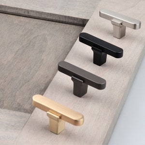 May include: Four cabinet pulls in different finishes: gold, nickel, black, and gunmetal. Each pull has a rectangular shape with rounded edges and a T-shaped design.