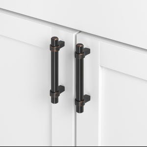 May include: Two black cabinet pulls with a copper finish. The pulls are mounted on white cabinet doors.