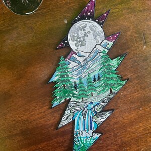 May include: An embroidered patch in the shape of a lightning bolt with a mountain scene, a full moon, and a waterfall. The patch is green, blue, white, and black.