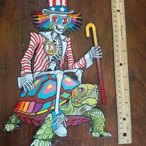 May include: An embroidered patch featuring a skeleton wearing a red, white, and blue top hat, sunglasses, and a striped jacket, riding a colorful turtle while holding a cane. A wooden ruler next to the patch shows a height of approximately 12 inches.