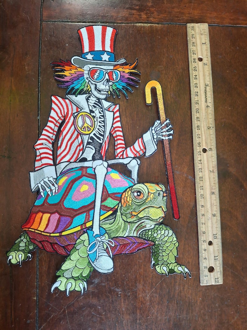 May include: An embroidered patch featuring a skeleton wearing a red, white, and blue top hat, sunglasses, and a striped jacket, riding a colorful turtle while holding a cane. A wooden ruler next to the patch shows a height of approximately 12 inches.