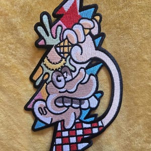 May include: An embroidered patch featuring a cartoon character with a tan face and large teeth, holding a waffle cone. The design includes a red and white checkered pattern at the base, a lightning bolt shape, and accents in blue, pink, and green.
