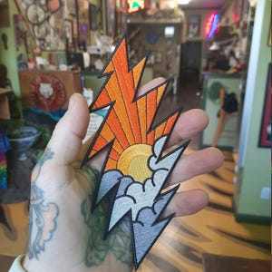 Könnte beinhalten: Ein farbenfroher gestickter Patch in Form eines Blitzes. Die obere Hälfte des Patches ist orange und gelb mit einer stilisierten Sonne, die über einem Gebirge aufgeht. Die untere Hälfte des Patches ist grau mit Wolken.