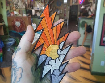 May include: A colorful embroidered patch in the shape of a lightning bolt. The top half of the patch is orange and yellow with a stylized sun rising over a mountain range. The bottom half of the patch is gray with clouds.