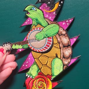 May include: Embroidered patch featuring a green turtle playing a banjo, with a pink and black lightning bolt design behind it. The turtle is standing on a red rose.