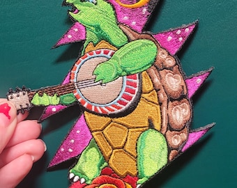 May include: Embroidered patch featuring a green turtle playing a banjo, with a pink and black lightning bolt design behind it. The turtle is standing on a red rose.