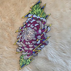 May include: Embroidered patch featuring a stylized chrysanthemum flower in shades of purple, gray, and white, set against a yellow lightning bolt design with green and blue accents. The patch is likely for clothing or accessories.