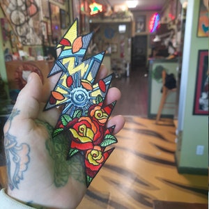 May include: A colorful embroidered patch featuring a lightning bolt design with a wheel, roses, and other details. The patch is held in a hand.