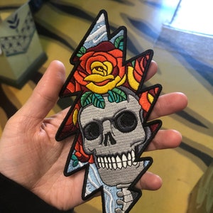 May include: An embroidered patch featuring a skull with a lightning bolt design. The skull is surrounded by colorful roses and has a blue and white background.