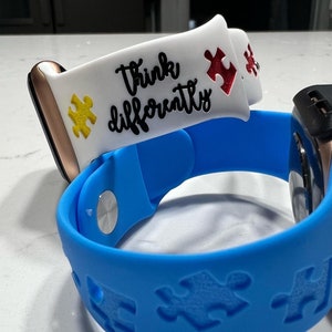 May include: Two smartwatch bands. One is blue with puzzle piece cutouts. The other is white with the words "Think Differently" and puzzle piece accents in yellow and red. The bands are attached to smartwatches.