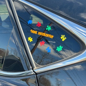 May include: A car window with colorful puzzle piece stickers and the words "THINK DIFFERENTLY" in orange lettering. The puzzle pieces are blue, red, green, and yellow. The car is black.