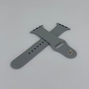 May include: Two gray silicone watch bands are displayed against a white background. One band is positioned horizontally, showing the holes for adjustment. The other band is crossed over it, showing the attachment mechanism.