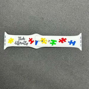 May include: White watch band with colorful puzzle pieces and the words "Think differently" in black script. The puzzle pieces are yellow, red, green, and blue. The band is designed for a smartwatch.