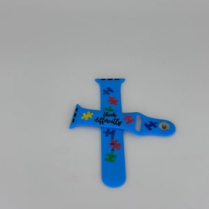 May include: Light blue smartwatch band with colorful puzzle piece graphics and the words "Think Differently." The band is made of a flexible material and features a stainless steel clasp. The band is designed to fit a smartwatch.