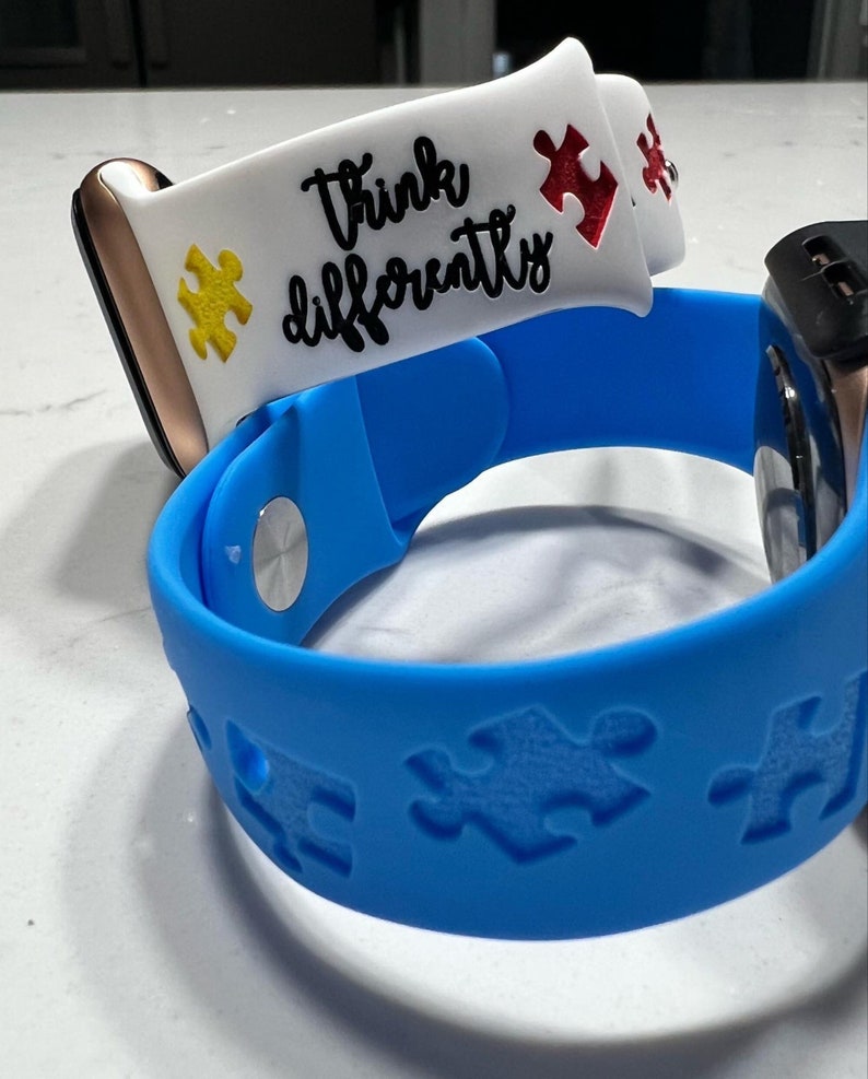 May include: Two smartwatch bands. One is blue with puzzle piece cutouts. The other is white with the words "Think Differently" and puzzle piece accents in yellow and red. The bands are attached to smartwatches.