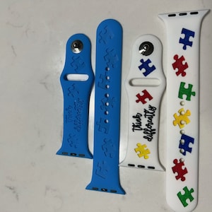 May include: Four colorful silicone watch bands. Two are blue, one is white. The blue bands have puzzle piece cutouts and the words "Think Differently". The white band has colorful puzzle pieces and the words "Think Differently".