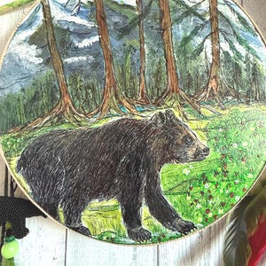 May include: A circular, hand-painted artwork showing a brown bear in a forest setting. The background features tall trees, mountains, and a green meadow with small red flowers. The artwork is framed with a gold-coloured trim.
