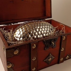 May include: A silver dragon egg with a textured surface sits inside a wooden treasure chest with brass accents and a hinged lid.