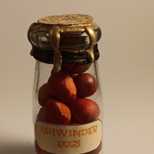 May include: A glass jar with a cork lid and a gold metal band. The jar is filled with red and orange Ashwinder eggs. The label on the jar reads "Ashwinder Eggs".