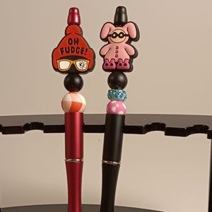May include: Two pens with decorative tops. The pen on the left has a red and white hat with the words "Oh Fudge!" and black glasses. The pen on the right has a pink bunny with black glasses and pink boots.