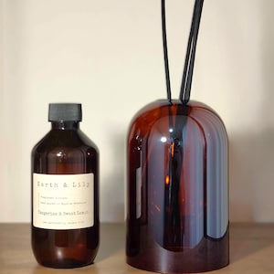 May include: A brown glass bottle with a black cap and a label reading Earth & Lily, Fragranced Diffuser, hand poured in Bayside, Melbourne, Tangerine & Sweet Lemon, next to a dome-shaped amber glass diffuser vessel with black reed sticks.