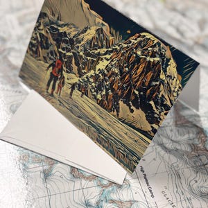 Puede incluir: Una tarjeta de felicitaci&oacute;n plegada con un paisaje monta&ntilde;oso estilizado con figuras, probablemente excursionistas. La tarjeta es blanca, con la imagen en tonos marrones, beige y azules. Descansa sobre un mapa topogr&aacute;fico detallado.