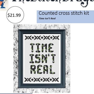 May include: Counted cross stitch kit featuring the phrase "Time Isn't Real" in a black frame. The kit includes a needle, white Aida fabric, hoop, thread, pattern, and instructions. Difficulty level is 2.