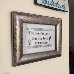 May include: A silver framed cross-stitch with the quote "The axe forgets but the tree remembers" in black lettering. A small tree is stitched in green below the quote.
