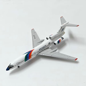 May include: A white model airplane featuring the Aeronica logo and text on the fuselage. The aircraft has a multi-colored stripe design in red, green, and blue near the nose and engines. The tail fin displays the registration code YN-CBT.