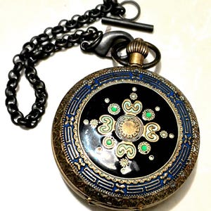 Op de afbeelding: Een antieke zakhorloge met een zwarte wijzerplaat, versierd met groene en witte bloemmotieven. Het horloge heeft een blauw-gouden rand en een zwarte ketting. De horlogekast is bronskleurig.