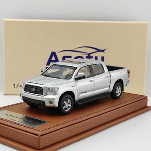 May include: A silver Toyota Tundra crew cab pickup truck scale model featuring a black grille, silver wheels, and a sunroof. The vehicle is displayed on a brown textured base with a metal plaque labelled Toyota Tundra Limited Edition.