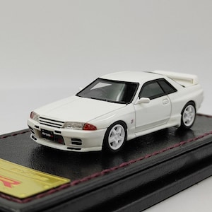 Peut inclure: Une maquette de voiture blanche avec des garnitures noires et des roues blanches. Le modèle est équipé d'un aileron et le mot "nismo" est visible à l'avant. La voiture est présentée sur une base noire.