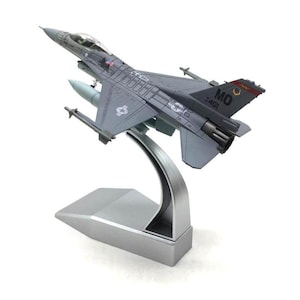 Peut inclure: Maquette d'avion de chasse F-16 grise, avec des marquages détaillés, montée sur un présentoir argenté. Le modèle présente un cockpit, des ailes et une queue détaillés, avec la queue marquée "MO 401". Le présentoir a une base rectangulaire et un support incurvé.