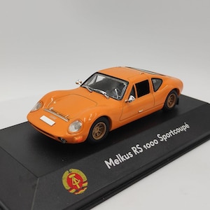 May include: A bright orange Melkus RS 1000 Sportcoupé scale model car mounted on a black display base. The base features the text Melkus RS 1000 Sportcoupé and a circular emblem with a hammer and compass design. The car has gold-colored wheels.