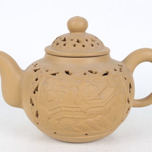 Può includere: Una teiera in ceramica marrone con un coperchio e un manico. La teiera ha intagli intricati e un disegno di uccelli e fiori. La teiera ha dei fori su tutto il corpo e il coperchio.
