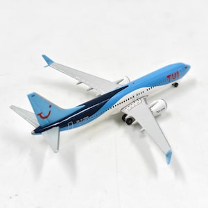 Puede incluir: Un modelo de avión con fuselaje azul y blanco, alas grises y el logotipo de TUI. La cola presenta un logotipo de cara sonriente. El avión tiene un diseño detallado, incluyendo motores y tren de aterrizaje. El texto "TUI.COM" es visible en el ala.