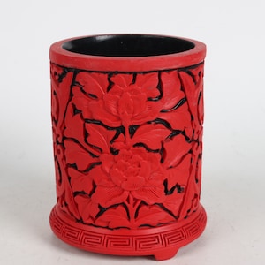 Puede incluir: Un recipiente cilíndrico rojo con un interior negro y tallas florales intrincadas. El recipiente tiene una base decorativa con un patrón geométrico repetitivo.