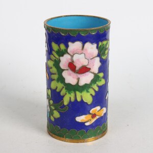 May include: A small, cylindrical blue enamelware cup with a floral design. The cup is decorated with pink, white, red, yellow, and green flowers and leaves. The cup has a gold rim.