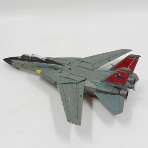 May include: A model of a gray and red F-14 Tomcat fighter jet. The model has detailed wings, tail fins, and a cockpit. The wings are swept back, and the tail fins are red with white and black markings. The model is on a white surface.