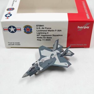 May include: A Lockheed Martin F-35A Lightning II model aircraft in a gray and white camouflage pattern, positioned in front of its white and red Herpa packaging box labeled with U.S. Air Force 65th Aggressor Squadron and Reg. 11-5020.