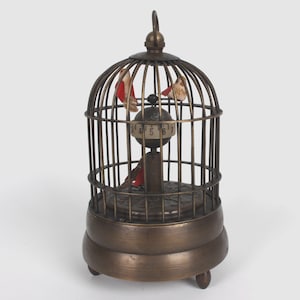 May include: A vintage-style brass birdcage clock with three small bird figurines inside. The clock face is a rotating sphere with numbers. The cage has a handle on top and sits on a round base with small feet. The birds are red and white.