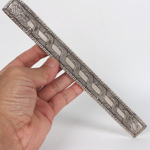 May include: A silver ruler featuring a decorative design of dragons and a repeating geometric pattern. The ruler is approximately 30 cm long.