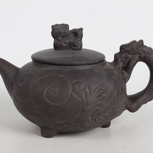 Può includere: Una teiera in ceramica marrone scuro con un manico a forma di drago e un coperchio con un piccolo animale scolpito. La teiera è decorata con intricate incisioni di motivi a spirale e una pianta stilizzata.