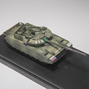 May include: A scale model of a green military tank featuring the number 211 on the turret, Cyrillic text on the hull, and small Russian flag decals on the sides. The tank has a long main gun barrel and detailed armor plating across the top surface.