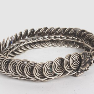 May include: A silver bracelet with a coiled design. The bracelet is made of many small, interconnected coils that form a continuous pattern. The bracelet has a clasp on one end.