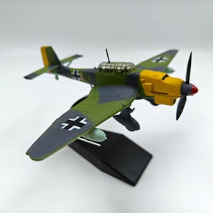May include: A detailed model airplane with a yellow, green, and gray color scheme. The model features a propeller, wings, and a cockpit. The wings have black and white markings. The model is mounted on a black stand.