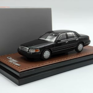 Könnte beinhalten: Ein schwarzes Modellauto einer Ford Crown Victoria Limousine auf einem braunen Sockel in Lederoptik mit schwarzer Plattform. Auf dem Sockel befindet sich der silberne Schriftzug Crown Victoria. Das Auto hat silberne Räder und klare Scheinwerfer.